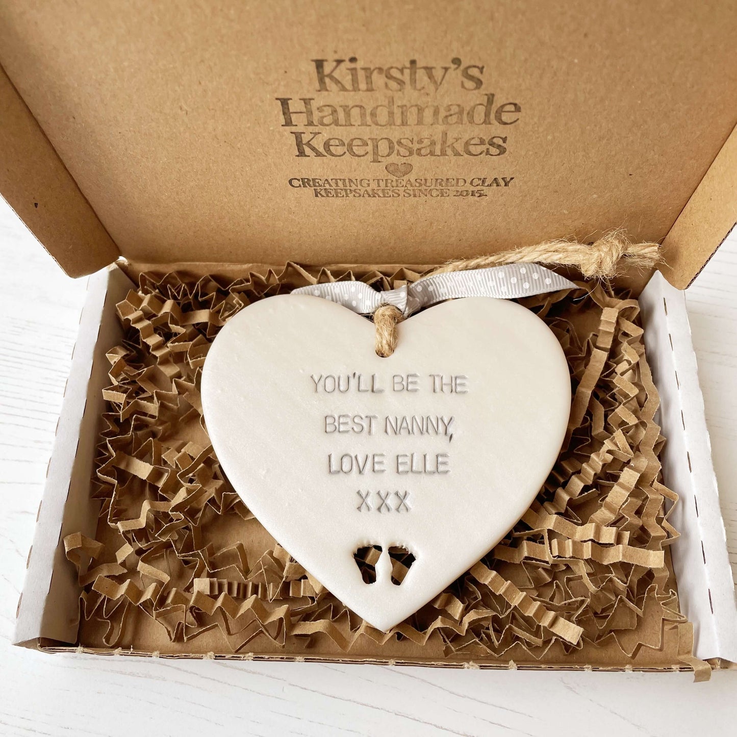 Pearlised white clay hanging heart pregnancy reveal with baby feet cut out of the bottom and grey personalisation, the heart is personalised with YOU’LL BE THE BEST NANNY, LOVE ELLE XXX