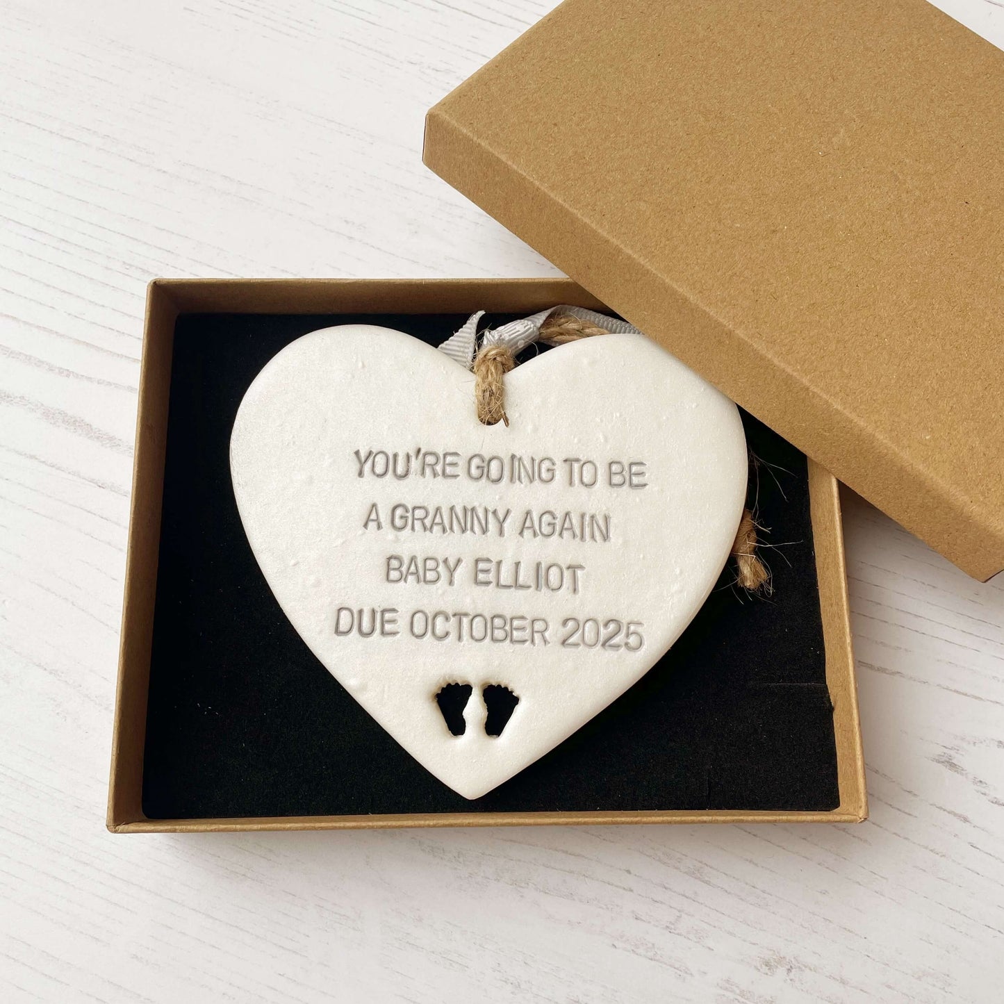 Pearlised white clay hanging heart with baby feet cut out of the bottom and grey personalisation, the heart is personalised with YOU’RE GOING TO BE A GRANNY AGAIN BABY ELLIOT DUE OCTOBER 2025