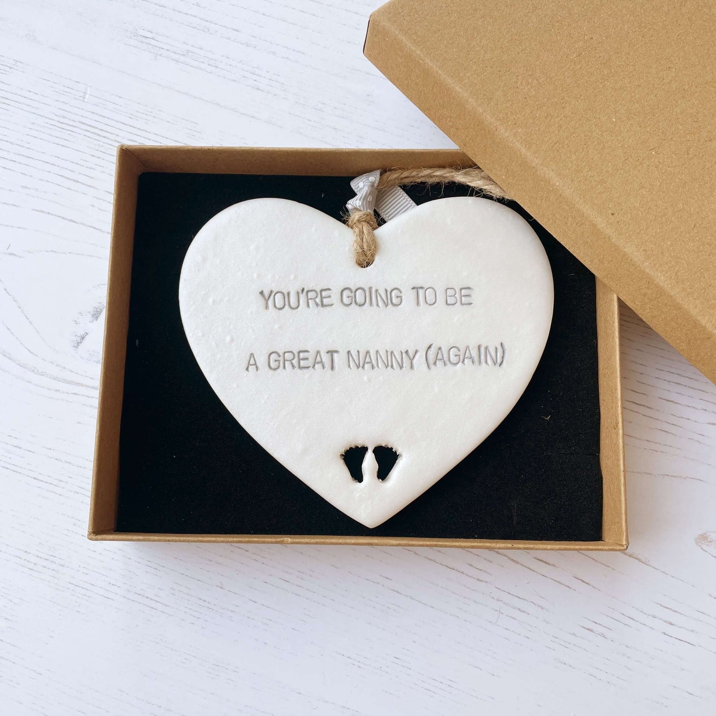 Pearlised white clay hanging heart with baby feet cut out of the bottom and grey personalisation, the heart is personalised with YOU’RE GOING TO BE GREAT NANNY (AGAIN)