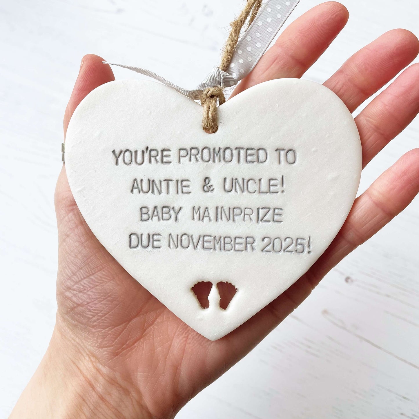 Pearlised white clay hanging heart with baby feet cut out of the bottom and grey personalisation, the heart is personalised with YOU’RE PROMOTED TO AUNTIE & UNCLE! BABY MAINPRIZE DUE NOVEMBER 2025!