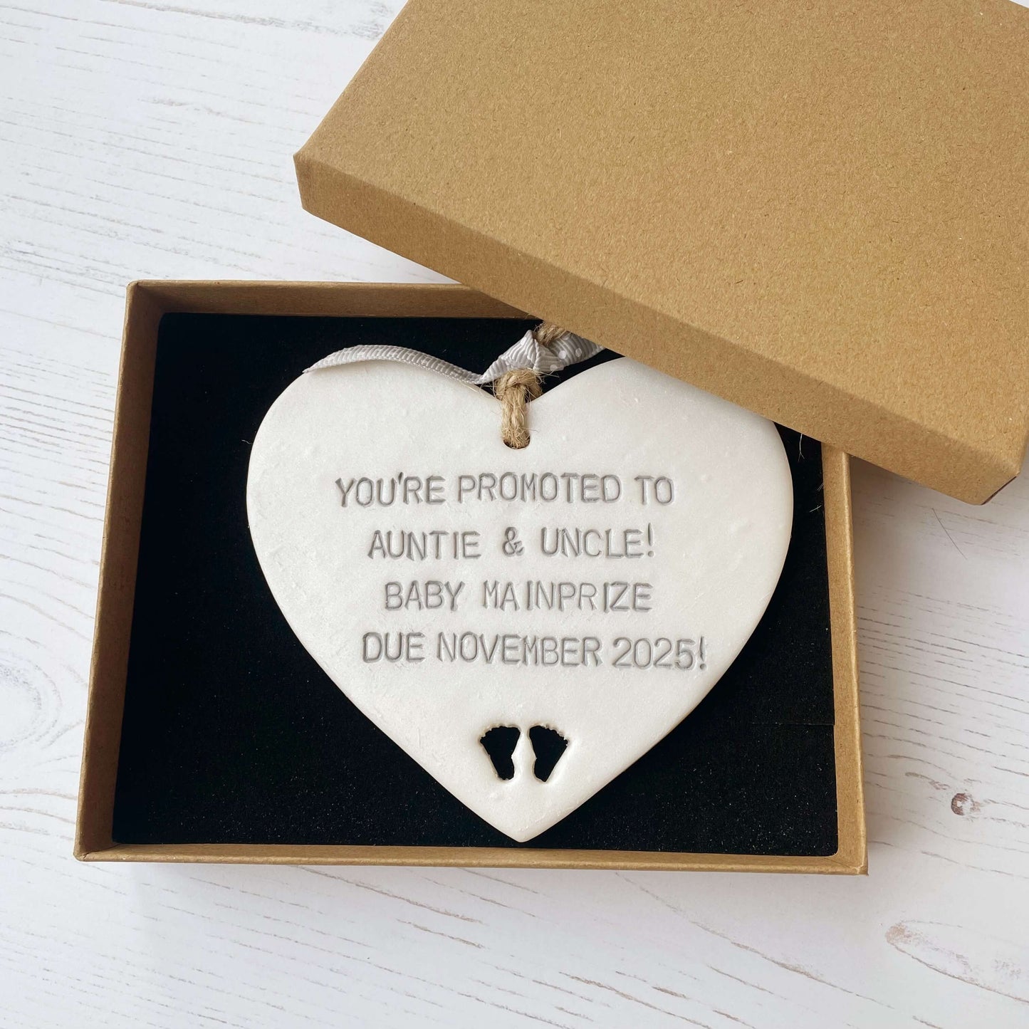 Pearlised white clay hanging heart with baby feet cut out of the bottom and grey personalisation, the heart is personalised with YOU’RE PROMOTED TO AUNTIE & UNCLE! BABY MAINPRIZE DUE NOVEMBER 2025!