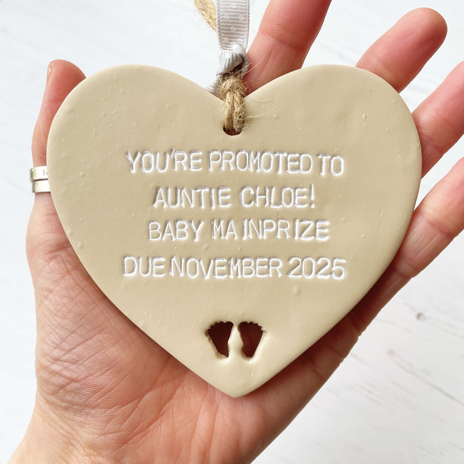 Beige clay hanging heart with baby feet cut out of the bottom and white personalisation, the heart is personalised with YOU’RE PROMOTED TO AUNTIE CHLOE! BABY MAINPRIZE DUE NOVEMBER 2025