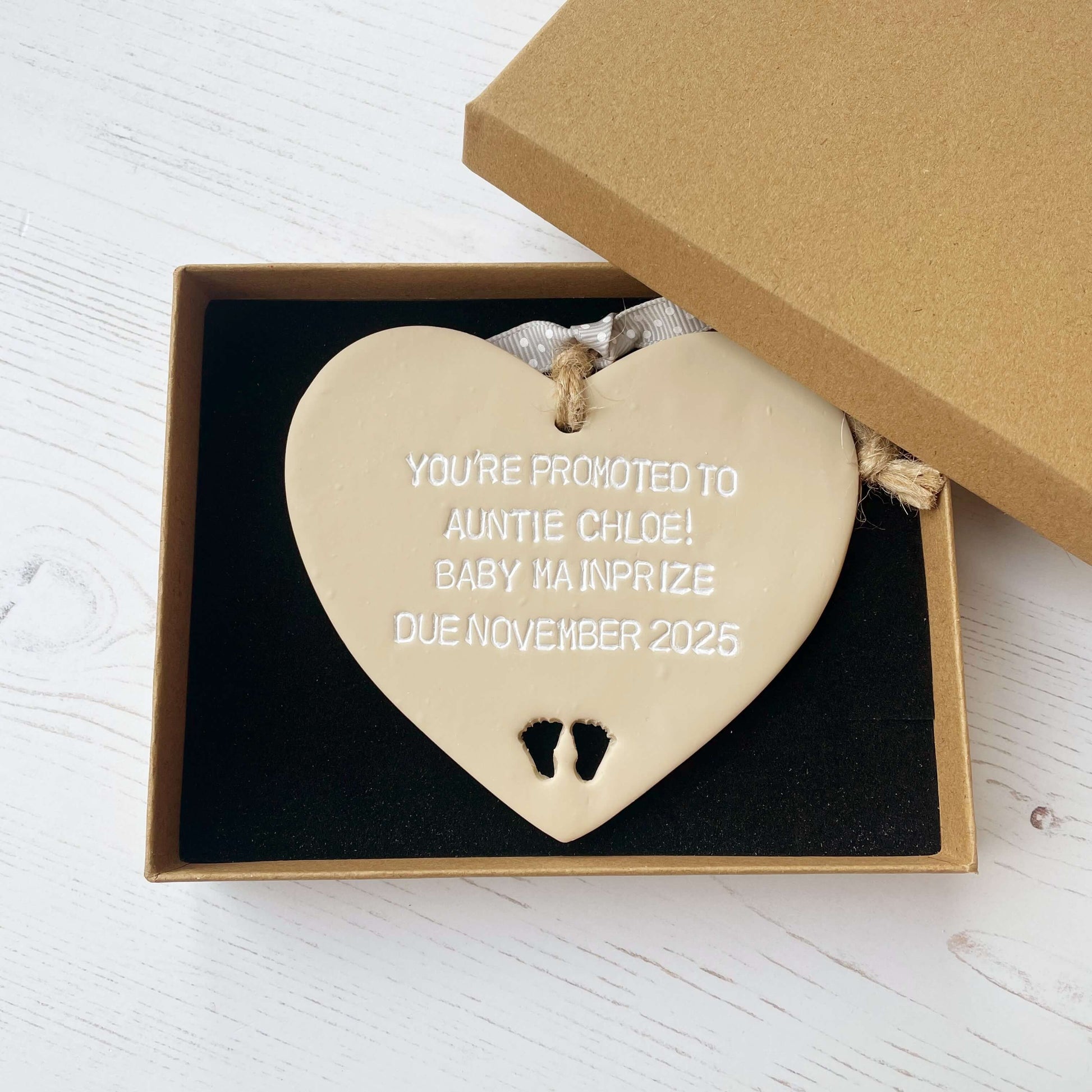 Beige clay hanging heart with baby feet cut out of the bottom and white personalisation, the heart is personalised with YOU’RE PROMOTED TO AUNTIE CHLOE! BABY MAINPRIZE DUE NOVEMBER 2025