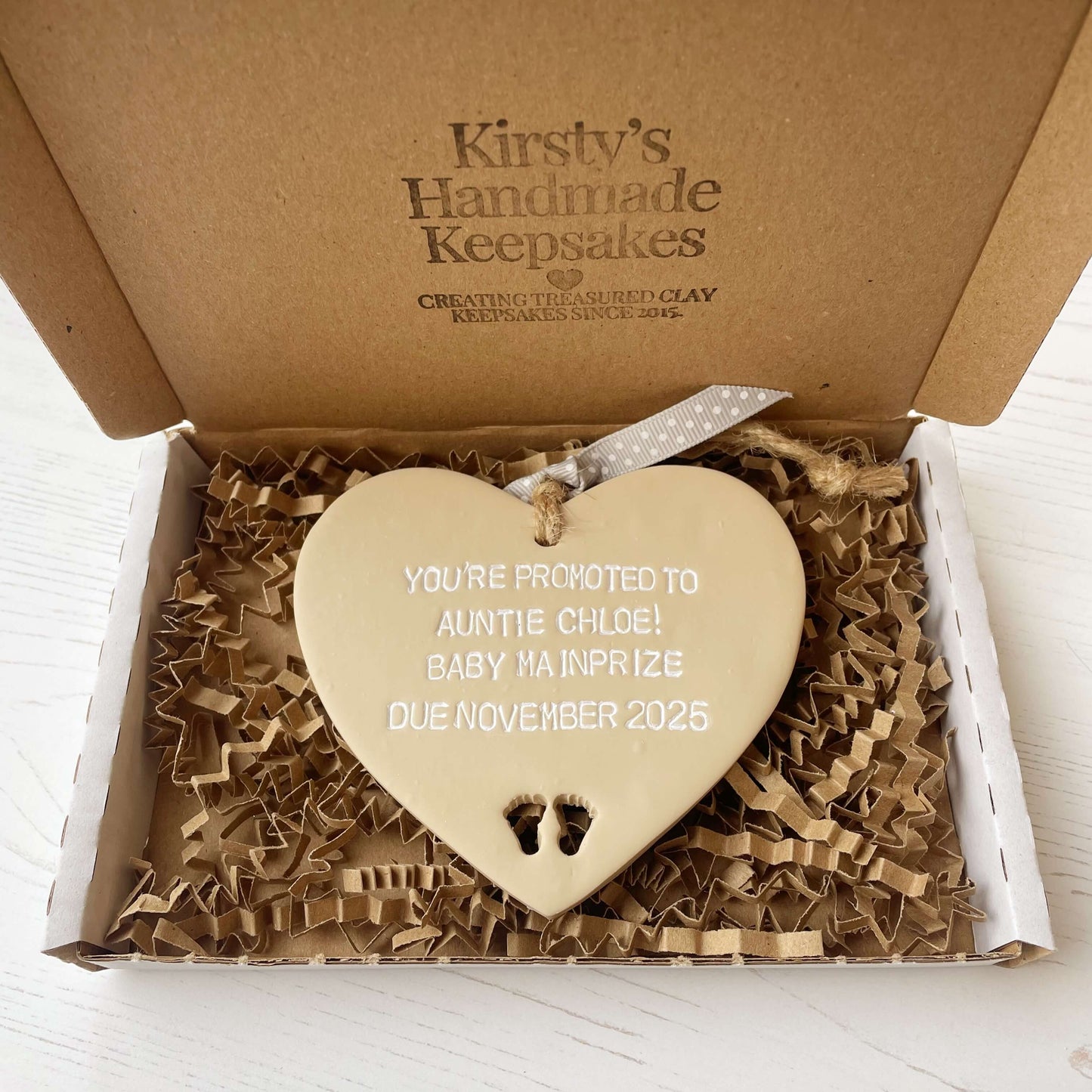 Beige clay hanging heart with baby feet cut out of the bottom and white personalisation, the heart is personalised with YOU’RE PROMOTED TO AUNTIE CHLOE! BABY MAINPRIZE DUE NOVEMBER 2025