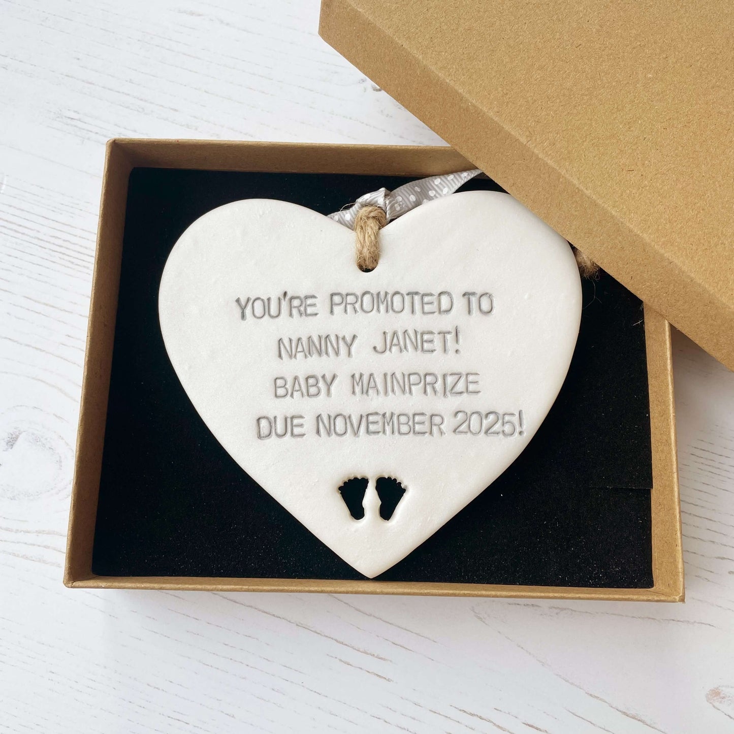 Pearlised white clay hanging heart with baby feet cut out of the bottom and grey personalisation, the heart is personalised with YOU’RE PROMOTED TO NANNY JANET! BABY MAINPRIZE DUE NOVEMBER 2025!