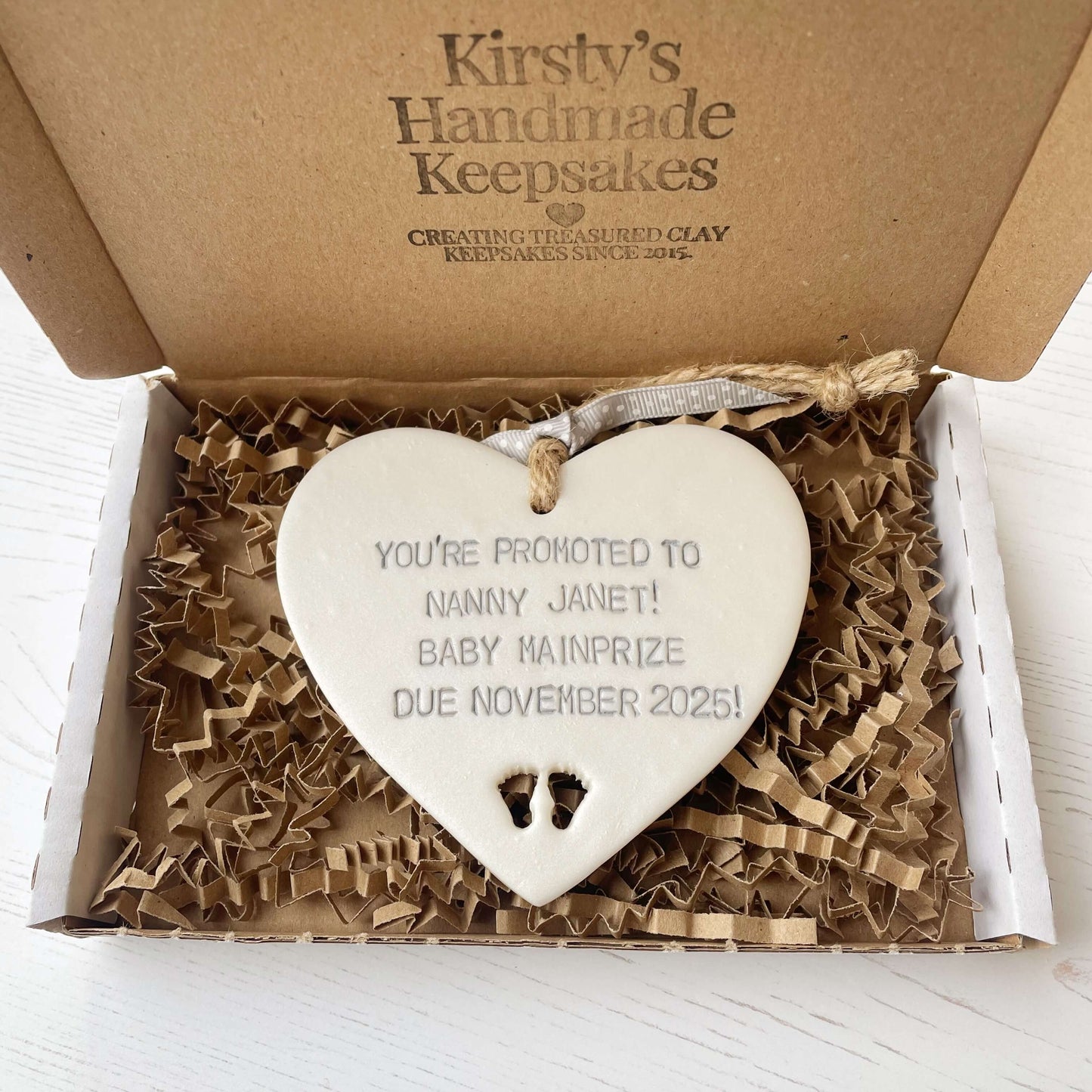 Pearlised white clay hanging heart with baby feet cut out of the bottom and grey personalisation, the heart is personalised with YOU’RE PROMOTED TO NANNY JANET! BABY MAINPRIZE DUE NOVEMBER 2025!
