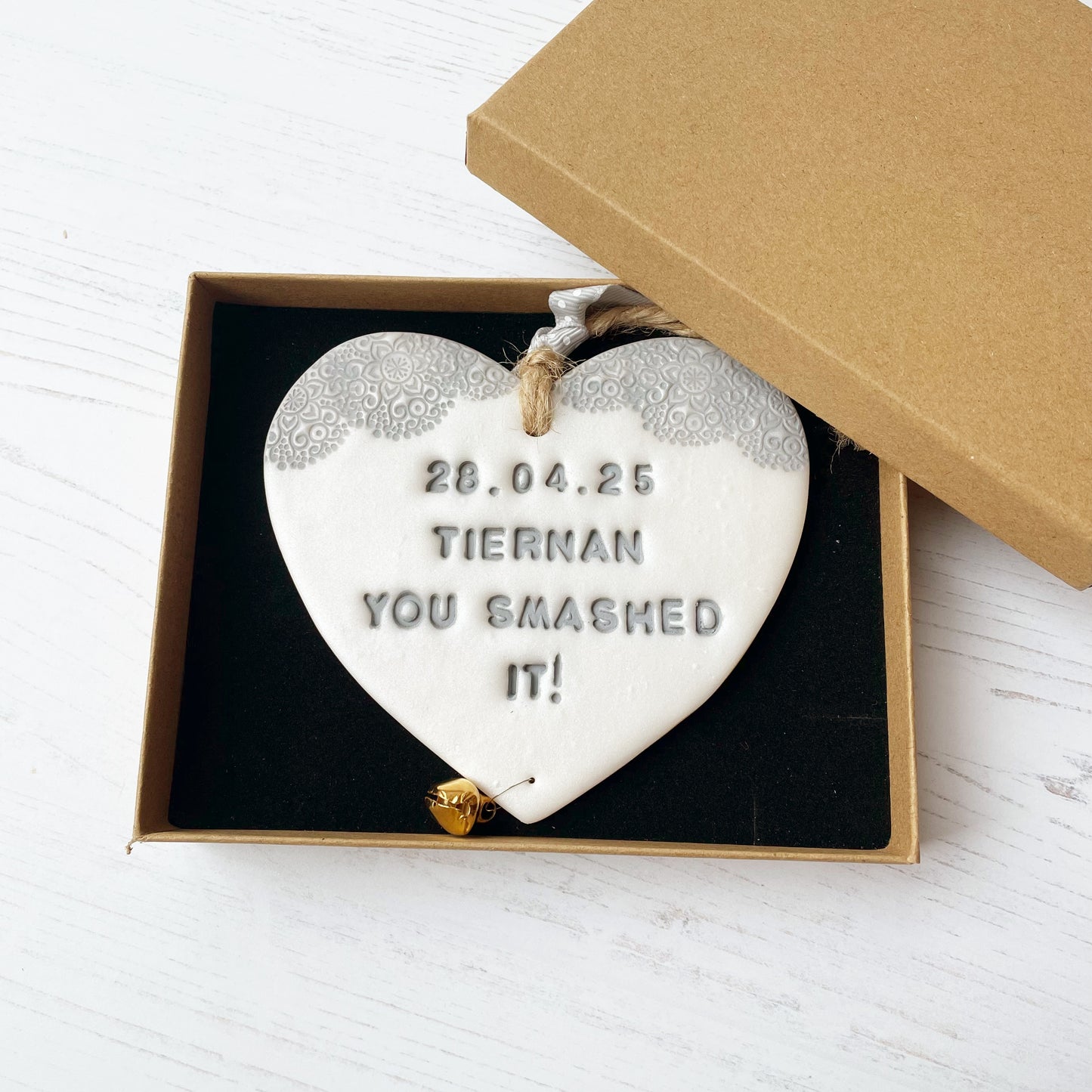 Personalised cancer survivor gift, pearlised white clay hanging heart with a grey lace edge at the top of the heart and a gold bell hanging below, the heart is personalised with 28.04.25 TIERNAN YOU SMASHED IT!