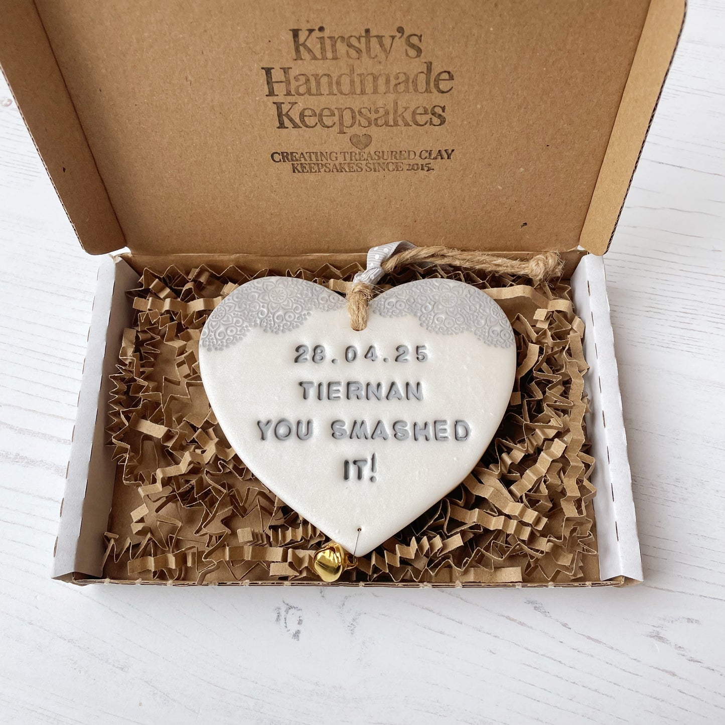 Personalised cancer survivor gift, pearlised white clay hanging heart with a grey lace edge at the top of the heart and a gold bell hanging below, the heart is personalised with 28.04.25 TIERNAN YOU SMASHED IT!