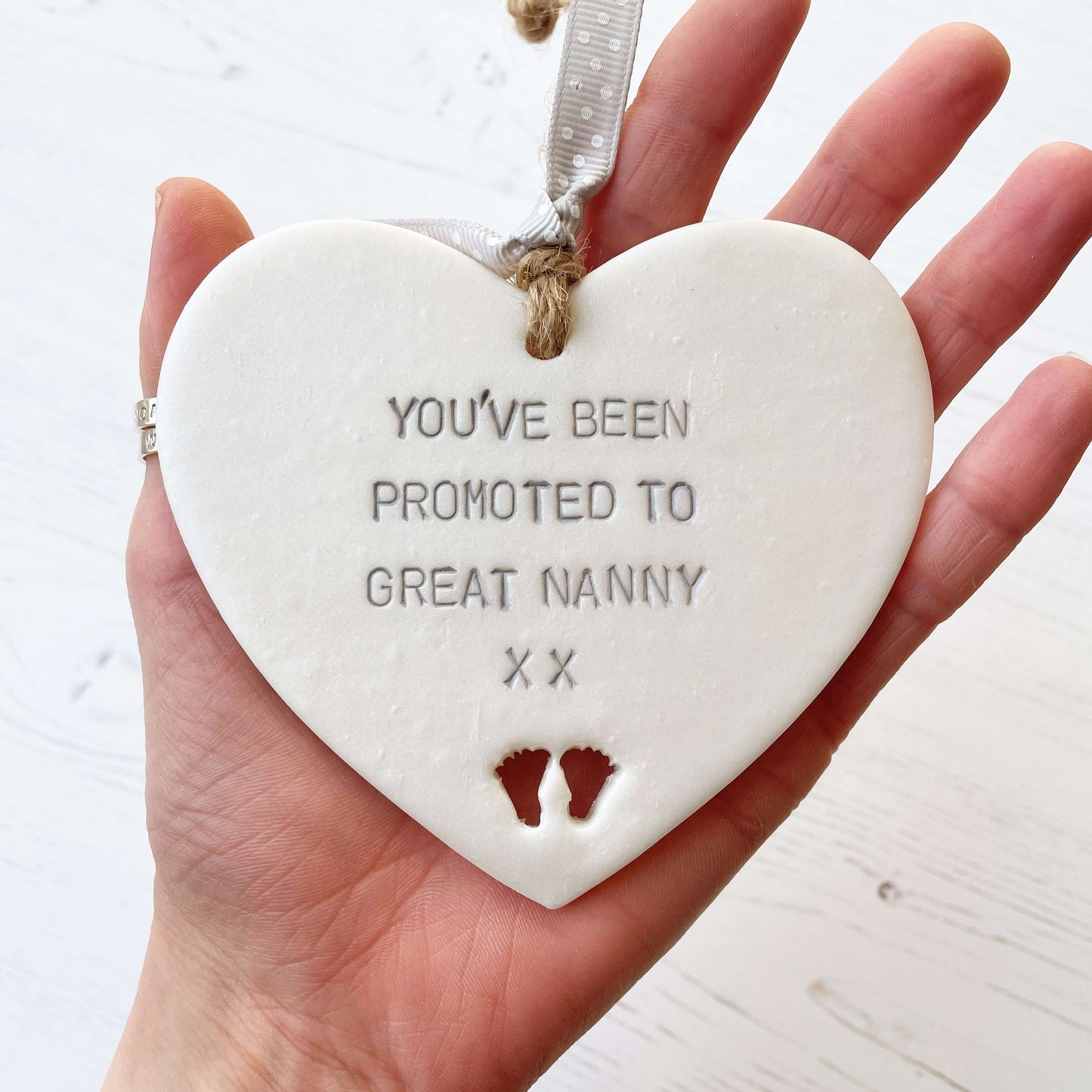 Pearlised white clay hanging heart with baby feet cut out of the bottom and grey personalisation, the heart is personalised with YOU’VE BEEN PROMOTED TO GREAT NANNY XX
