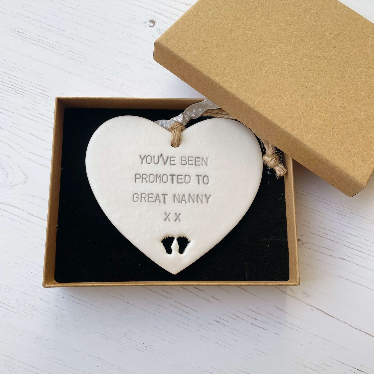 Pearlised white clay hanging heart with baby feet cut out of the bottom and grey personalisation, the heart is personalised with YOU’VE BEEN PROMOTED TO GREAT NANNY XX