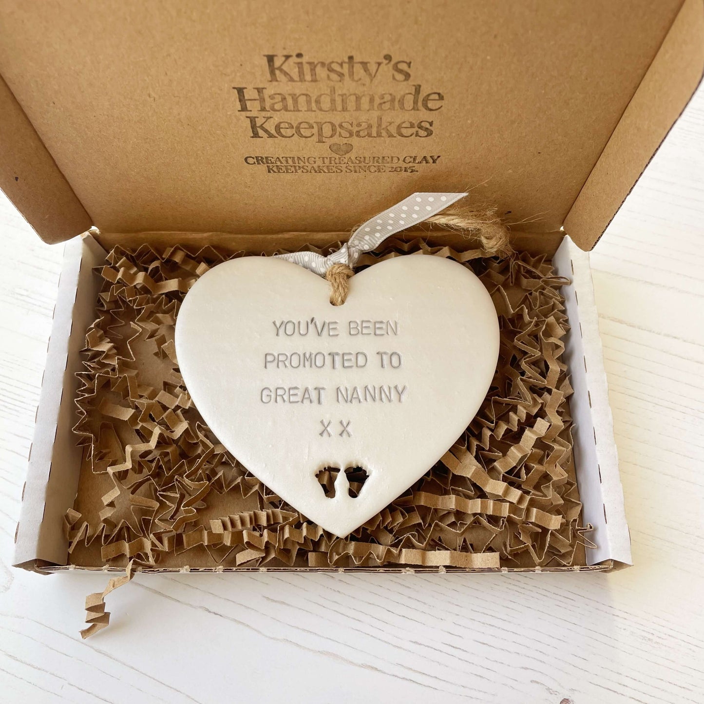 Pearlised white clay hanging heart with baby feet cut out of the bottom and grey personalisation, the heart is personalised with YOU’VE BEEN PROMOTED TO GREAT NANNY XX
