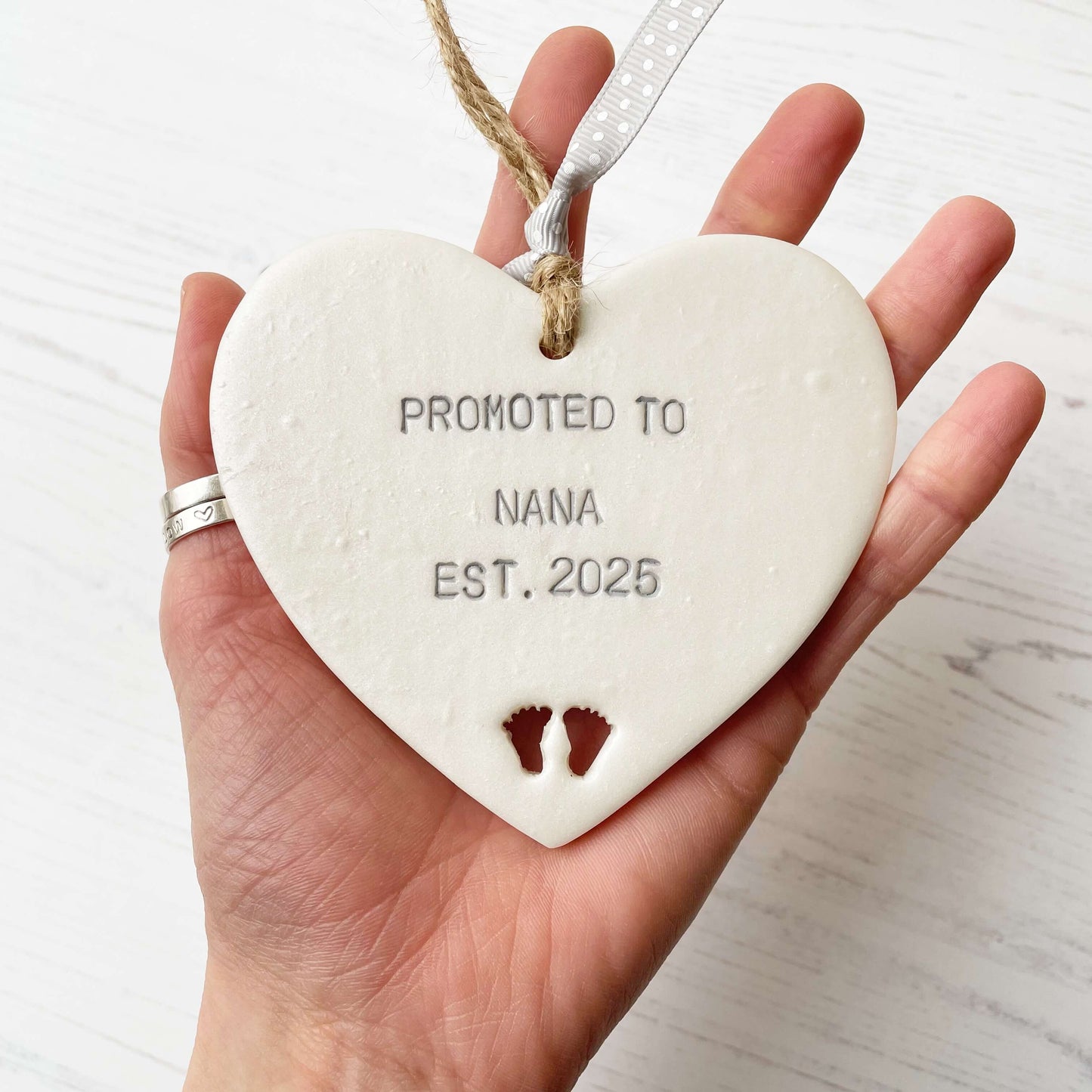 Pearlised white clay hanging heart with baby feet cut out of the bottom and grey personalisation, the heart is personalised with PROMOTED TO NANA EST. 2025