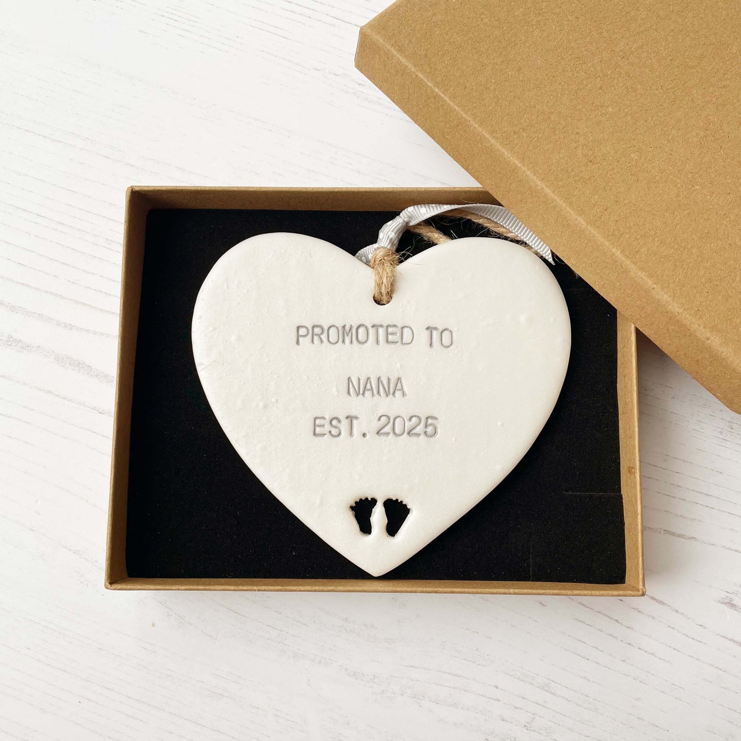 Pearlised white clay hanging heart with baby feet cut out of the bottom and grey personalisation, the heart is personalised with PROMOTED TO NANA EST. 2025