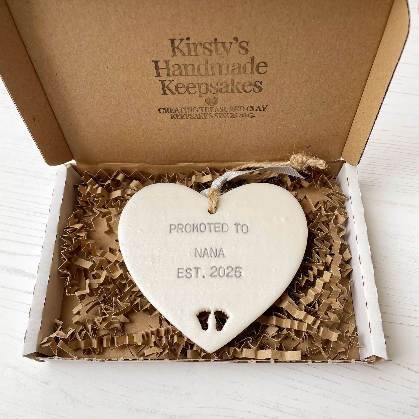 Pearlised white clay hanging heart with baby feet cut out of the bottom and grey personalisation, the heart is personalised with PROMOTED TO NANA EST. 2025