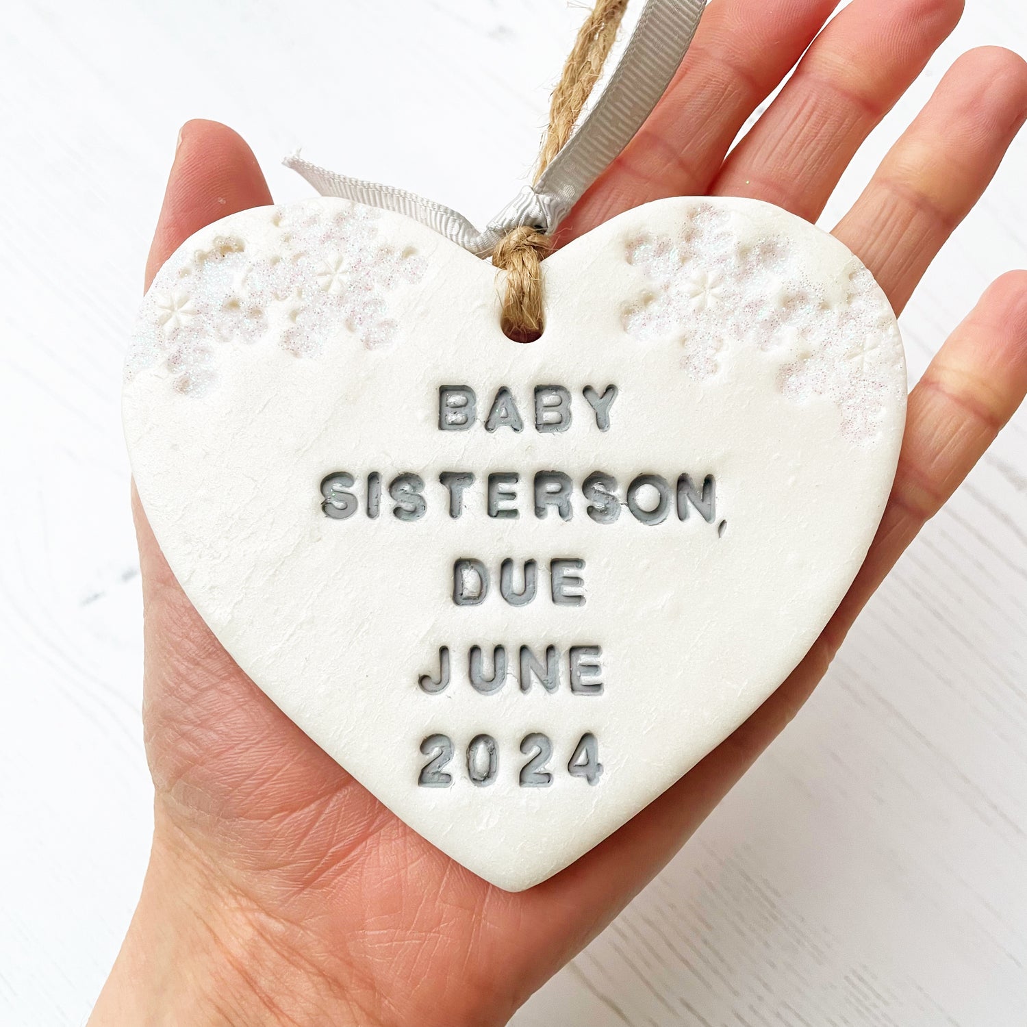 Personalised baby's announcement Christmas heart ornament, pearlised white clay with BABY SISTERSON, DUE JUNE 2024 painted grey, decorated with 2 iridescent glitter snowflakes on either side of the top of the heart