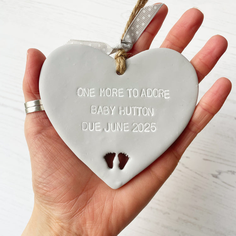 Grey clay hanging heart with baby feet cut out of the bottom and white personalisation, the heart is personalised with ONE MORE TO ADORE BABY HUTTON DUE JUNE 2025