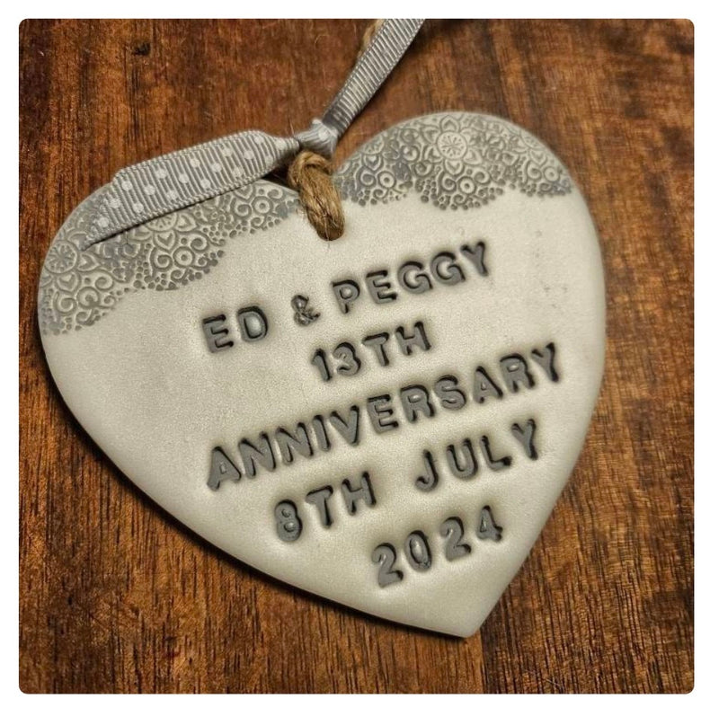 A Pearlised white polymer clay heart anniversary ornament hand stamped with 'ED & PEGGY 13TH ANNIVERSARY 8TH JULY 2024', hanging from a silver ribbon on a rustic wooden background.