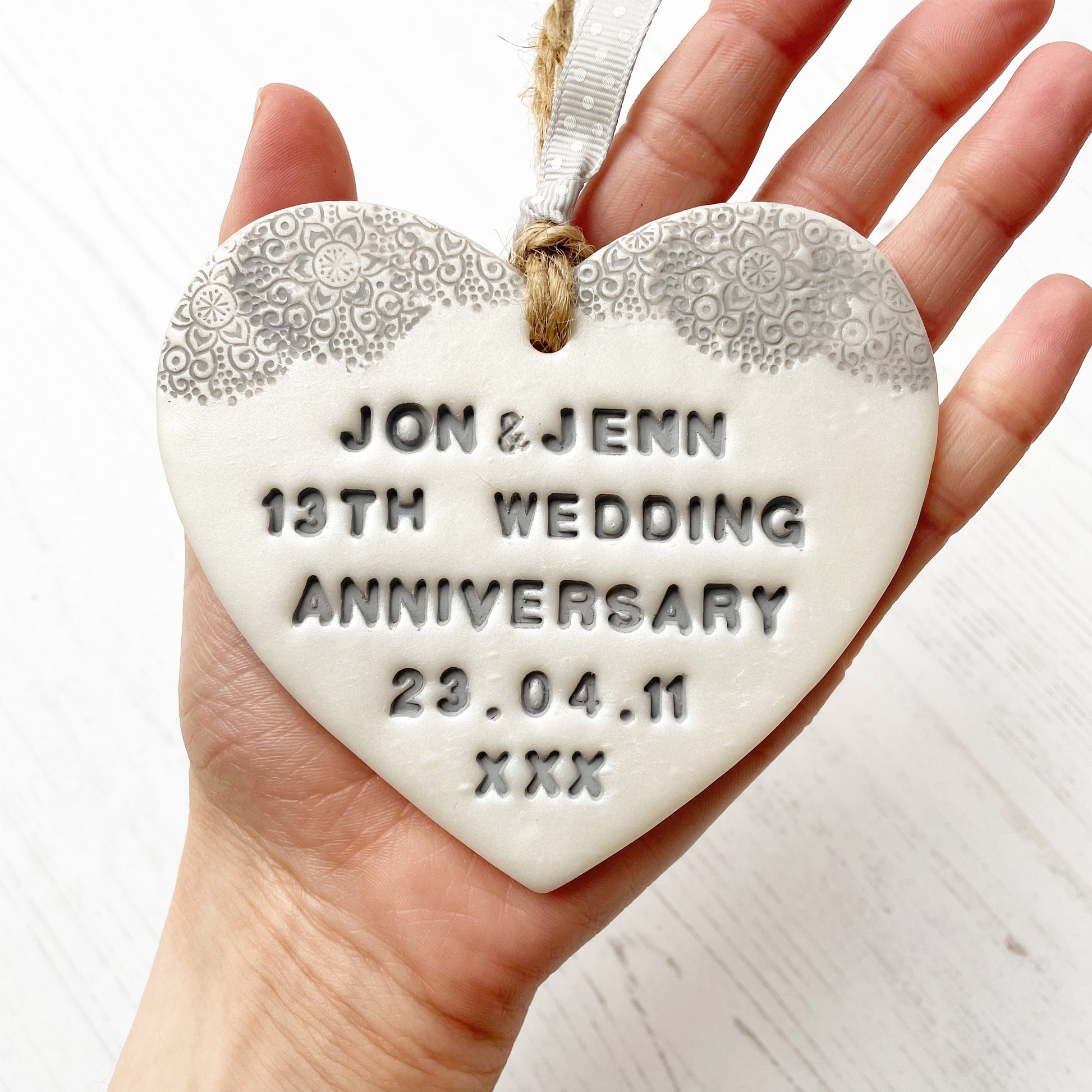 Personalised 13th anniversary gift, pearlised white clay hanging heart with a grey lace edge at the top of the heart, the heart is personalised with JON & JENN 13TH WEDDING ANNIVERSARY 23.04.11 XXX