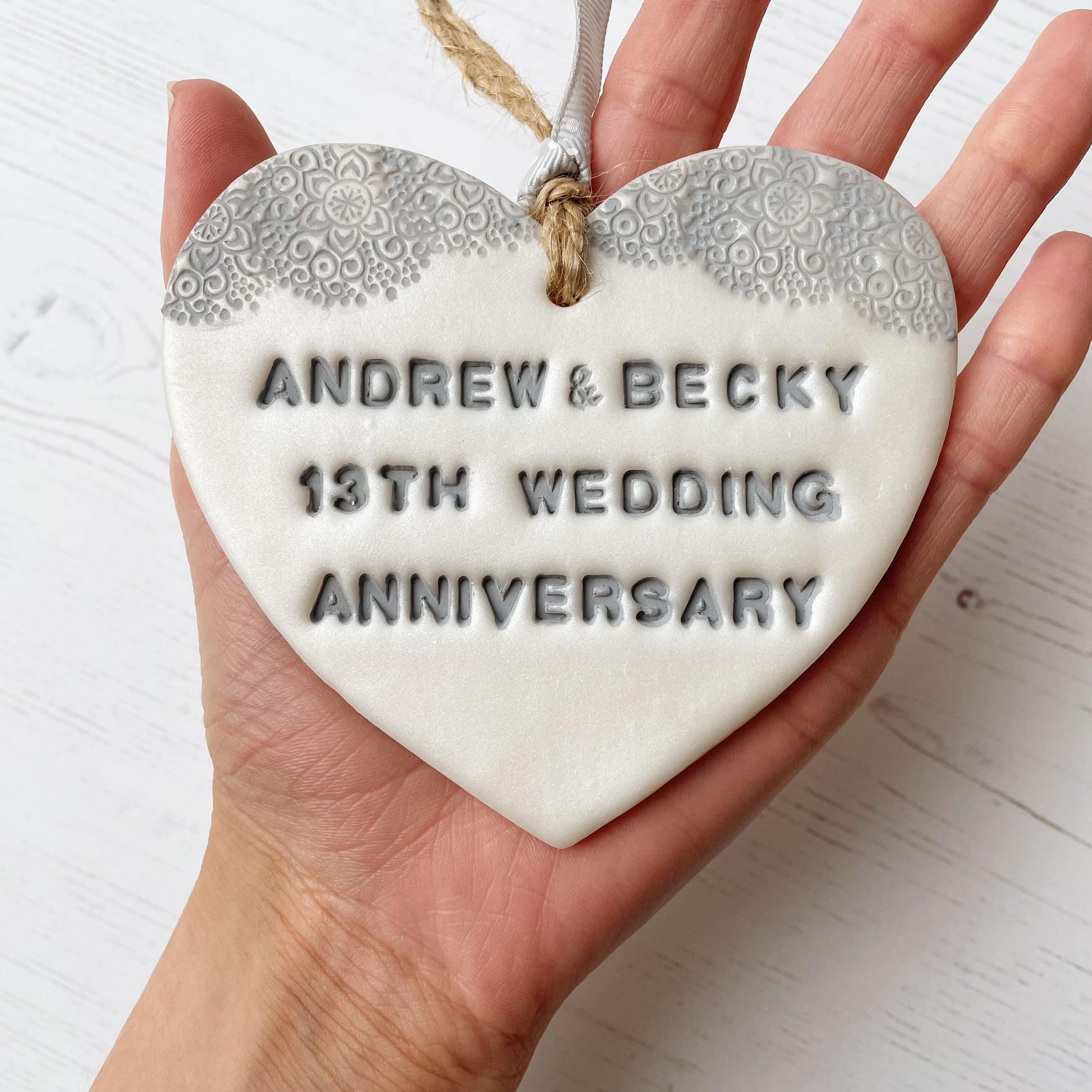 Personalised 13th anniversary gift, pearlised white clay hanging heart with a grey lace edge at the top of the heart, the heart is personalised with ANDREW & BECKY 13TH WEDDING ANNIVERSARY