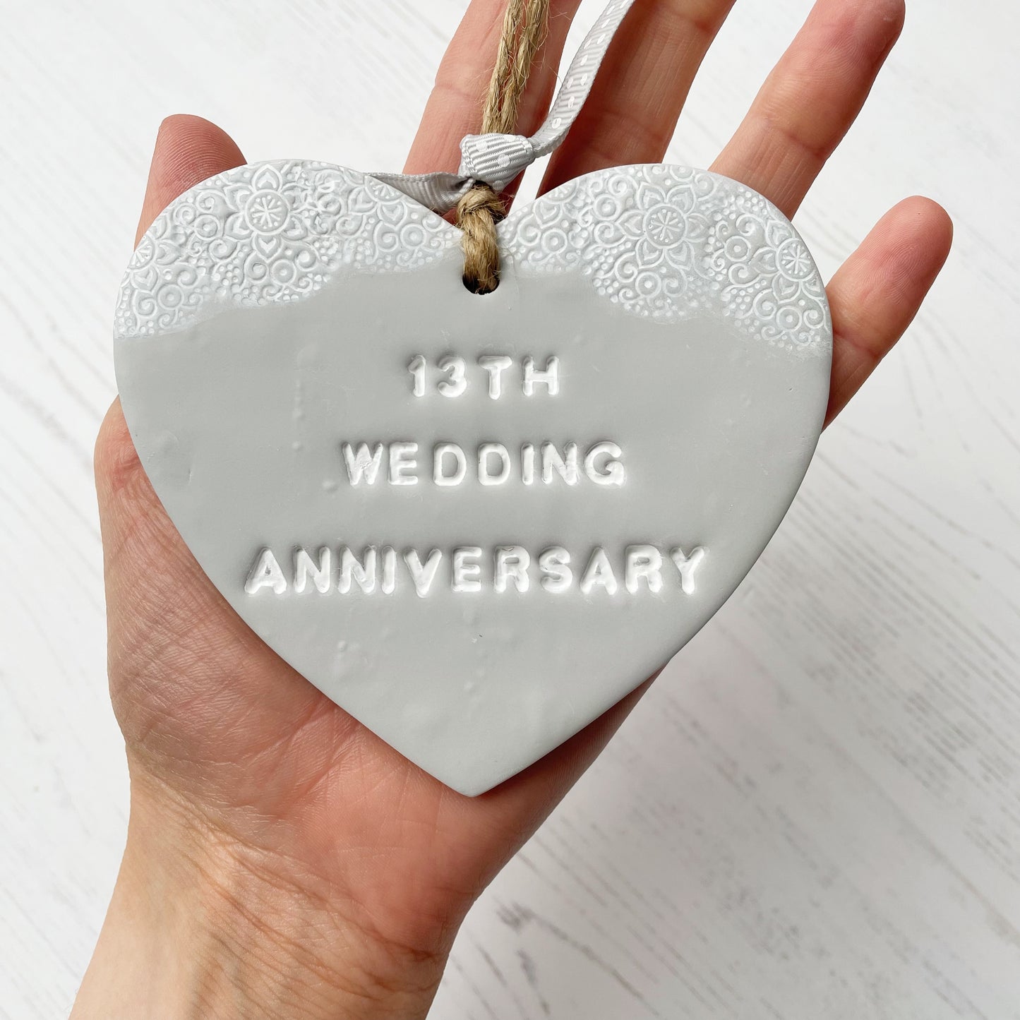 13th wedding anniversary gift, grey clay heart with a white lace edge at the top of the heart with jute twine for hanging, the heart is personalised with 13TH WEDDING ANNIVERSARY
