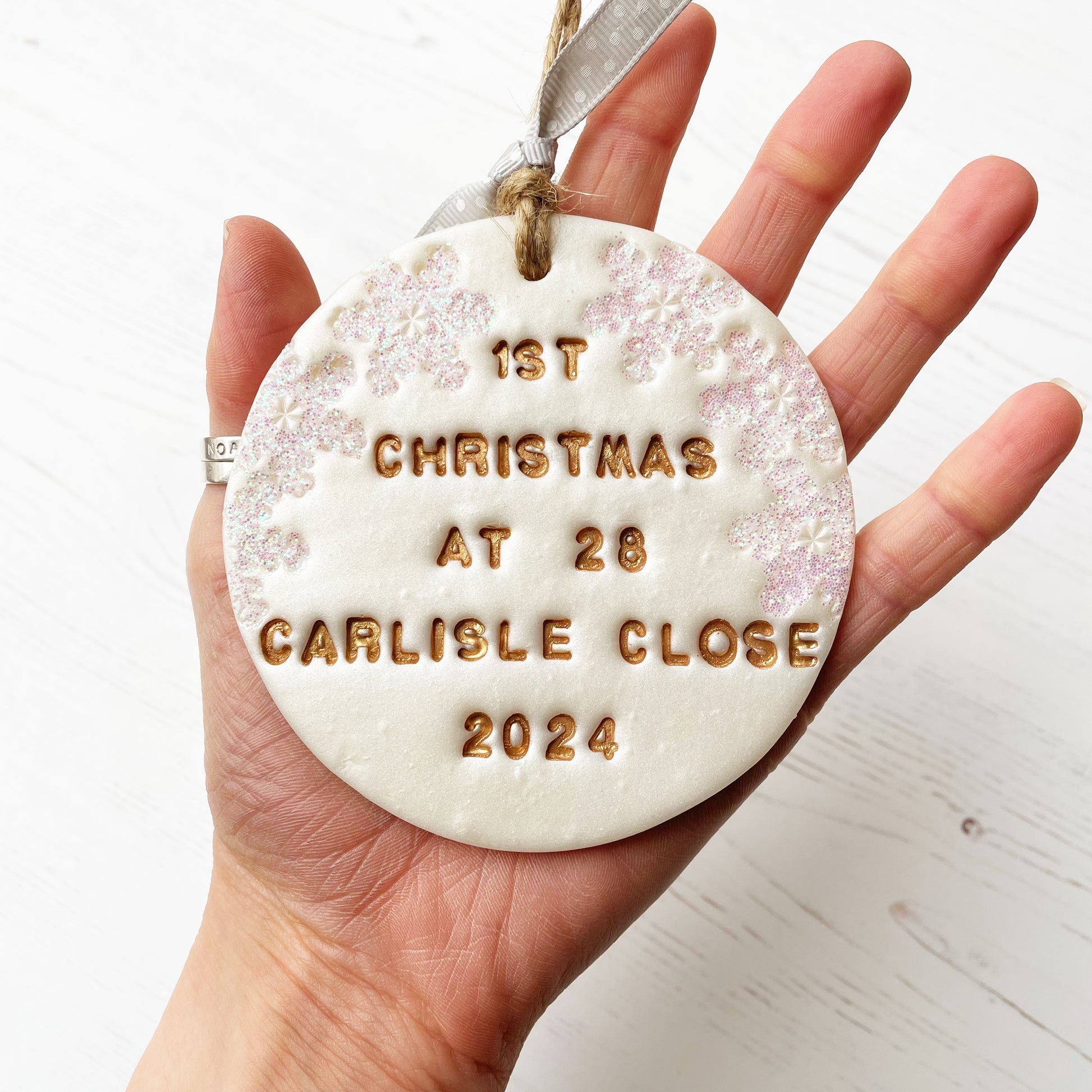 Personalised first Christmas in our new home bauble ornament, pearlised white round clay with 1ST CHRISTMAS AT 28 CARLISLE CLOSE 2024 in gold paint, decorated with 3 iridescent glitter snowflakes on either side of the bauble