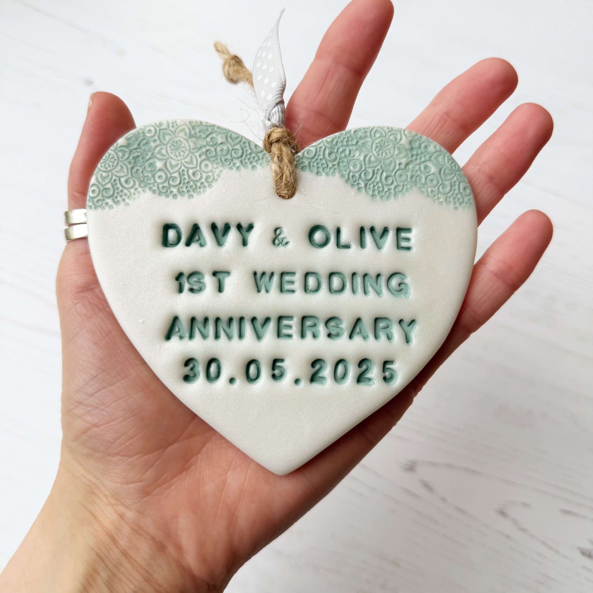 Personalised 1st wedding anniversary gift, pearlised white clay hanging heart with a sage green lace edge at the top of the heart, the heart is personalised with DAVY & OLIVE 1ST WEDDING ANNIVERSARY 30.05.2025