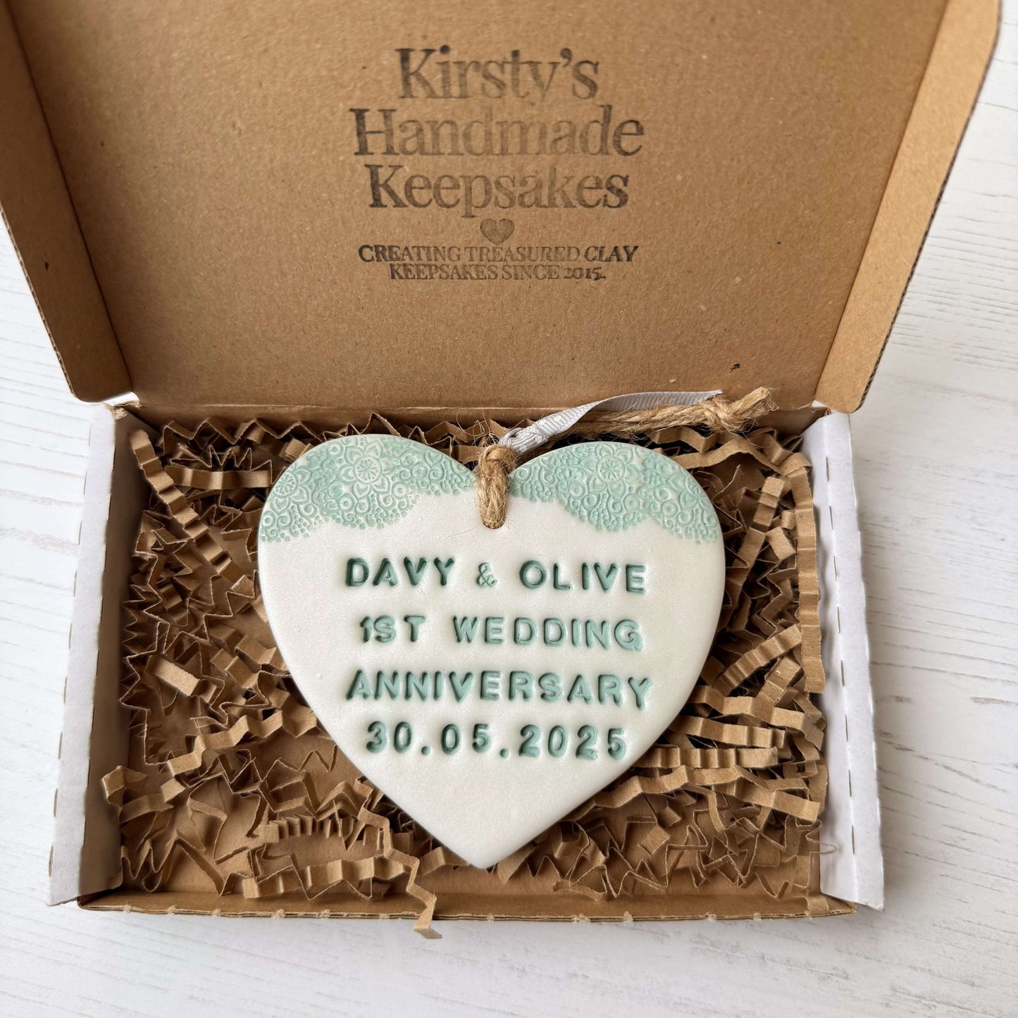 Personalised 1st wedding anniversary gift, pearlised white clay hanging heart with a sage green lace edge at the top of the heart, the heart is personalised with DAVY & OLIVE 1ST WEDDING ANNIVERSARY 30.05.2025