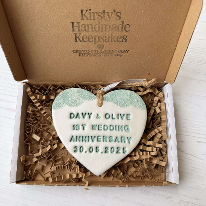 Personalised 1st wedding anniversary gift, pearlised white clay hanging heart with a sage green lace edge at the top of the heart, the heart is personalised with DAVY & OLIVE 1ST WEDDING ANNIVERSARY 30.05.2025
