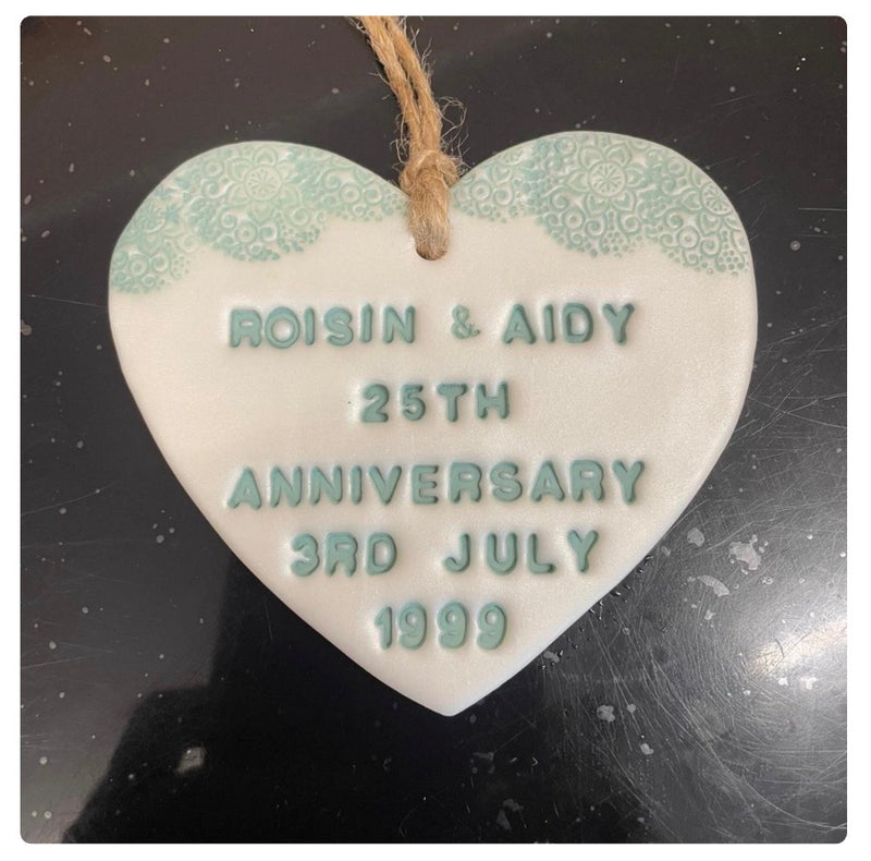A Pearlised white polymer clay heart  anniversary ornament with a natural jute hanging string, featuring embossed green text reading 'ROISIN & AIDY 25TH ANNIVERSARY 3RD JULY 1999' on a white background with a light green floral pattern at the top corners.