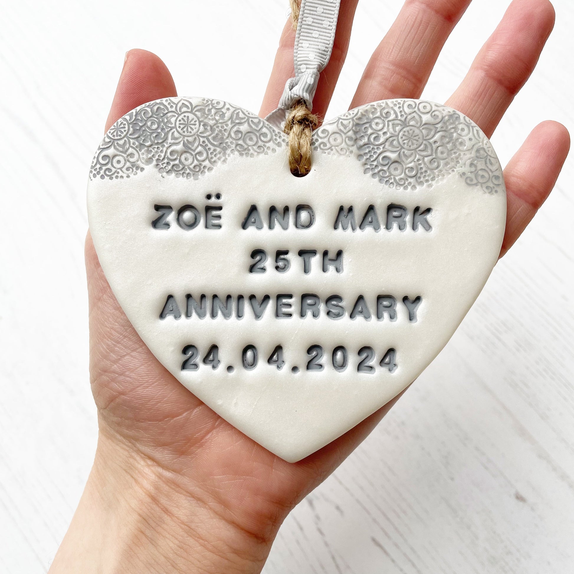 Personalised 25th anniversary gift, pearlised white clay hanging heart with a grey lace edge at the top of the heart, the heart is personalised with ZOË AND MARK 25TH ANNIVERSARY 24.04.2024