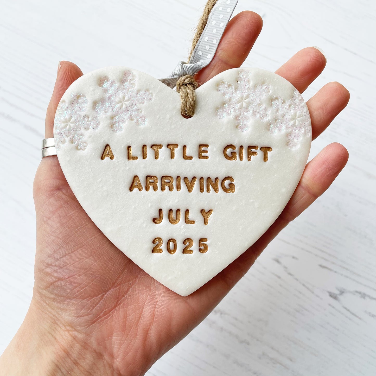 Personalised baby reveal Christmas heart ornament, pearlised white clay with A LITTLE GIFT ARRIVING JULY 2025 painted gold, decorated with 2 iridescent glitter snowflakes on either side of the top of the heart