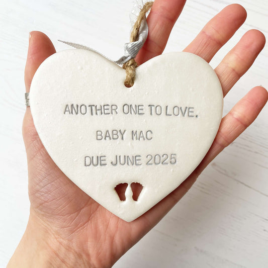 Pearlised white clay hanging heart with baby feet cut out of the bottom and grey personalisation, the heart is personalised with ANOTHER ONE TO LOVE. BABY MAC DUE JUNE 2025