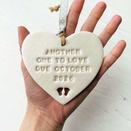Personalised baby reveal gift, pearlised white clay heart with feet cut out of the bottom personalised with ANOTHER ONE TO LOVE DUE OCTOBER 2026