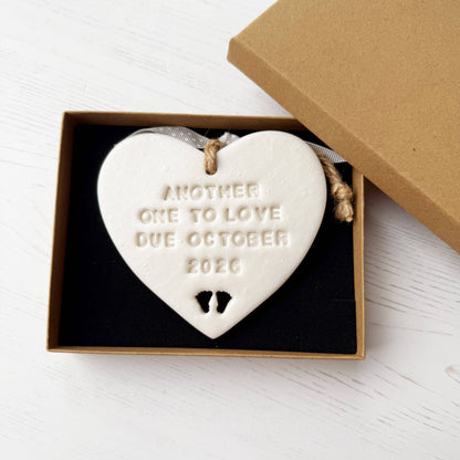 Personalised baby reveal gift, pearlised white clay heart with feet cut out of the bottom personalised with ANOTHER ONE TO LOVE DUE OCTOBER 2026