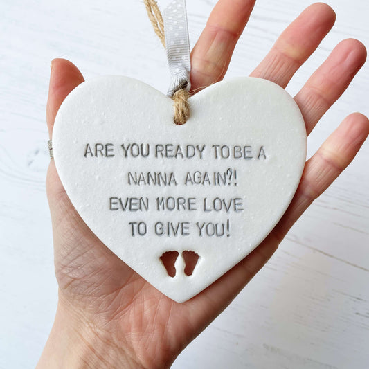 Pearlised white clay hanging heart pregnancy reveal with baby feet cut out of the bottom and grey personalisation, the heart is personalised with ARE YOU READY TO BE A NANNA, AGAIN?! EVEN MORE LOVE TO GIVE YOU!
