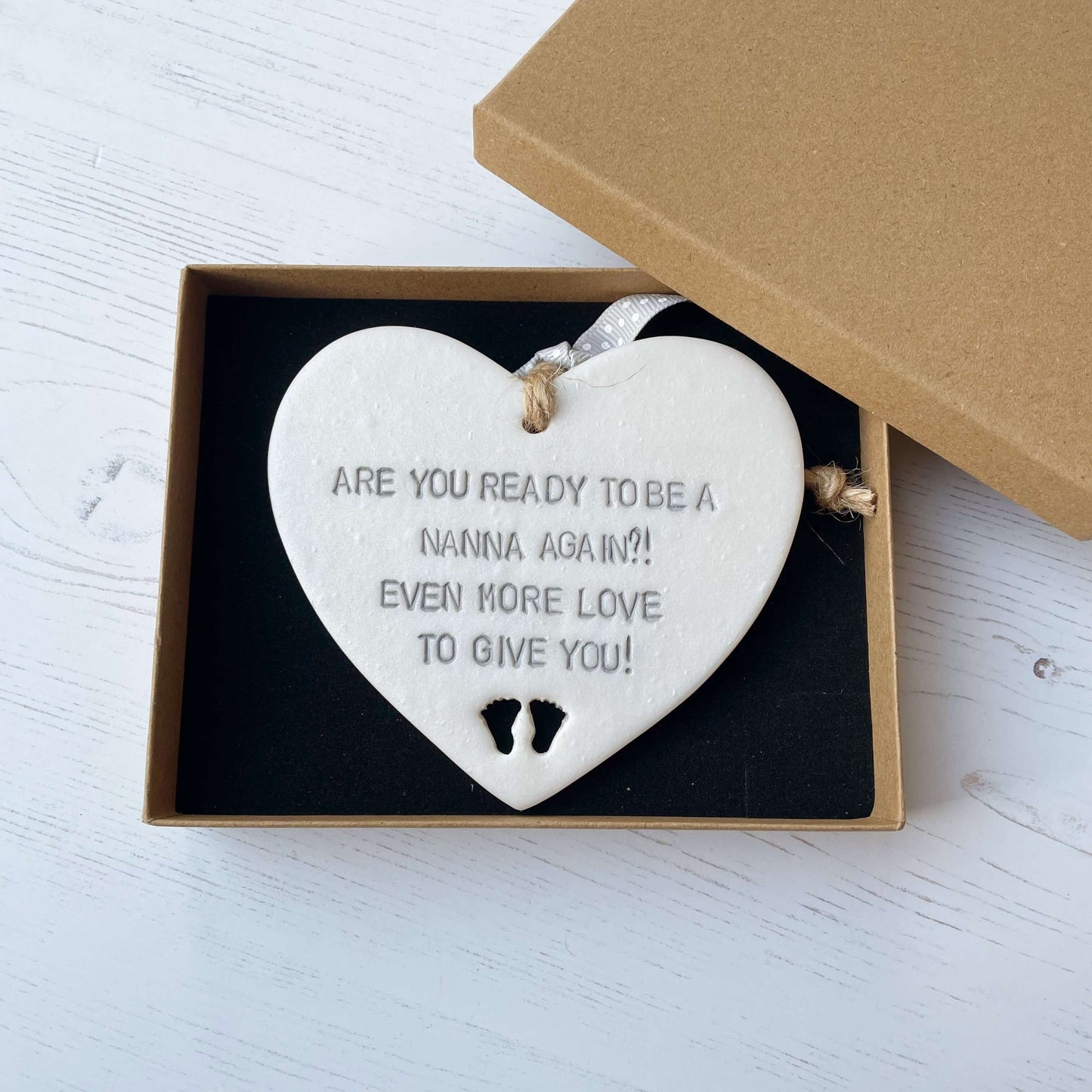 Pearlised white clay hanging heart pregnancy reveal with baby feet cut out of the bottom and grey personalisation, the heart is personalised with ARE YOU READY TO BE A NANNA, AGAIN?! EVEN MORE LOVE TO GIVE YOU!