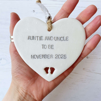 Pearlised white clay hanging heart with baby feet cut out of the bottom and grey personalisation, the heart is personalised with AUNTIE AND UNCLE TO BE NOVEMBER 2025