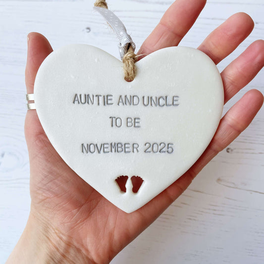 Pearlised white clay hanging heart with baby feet cut out of the bottom and grey personalisation, the heart is personalised with AUNTIE AND UNCLE TO BE NOVEMBER 2025