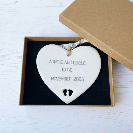Pearlised white clay hanging heart with baby feet cut out of the bottom and grey personalisation, the heart is personalised with AUNTIE AND UNCLE TO BE NOVEMBER 2025
