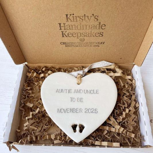 Pearlised white clay hanging heart with baby feet cut out of the bottom and grey personalisation, the heart is personalised with AUNTIE AND UNCLE TO BE NOVEMBER 2025