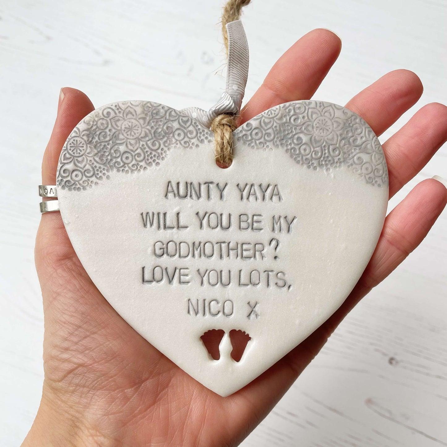 Personalised Godmother gift, pearlised white clay hanging heart with baby feet cut out of the bottom and grey lace edge at the top of the heart, the heart is personalised with AUNTY YAYA, WILL YOU BE MY GODMOTHER? LOVE YOU LOTS NICO X