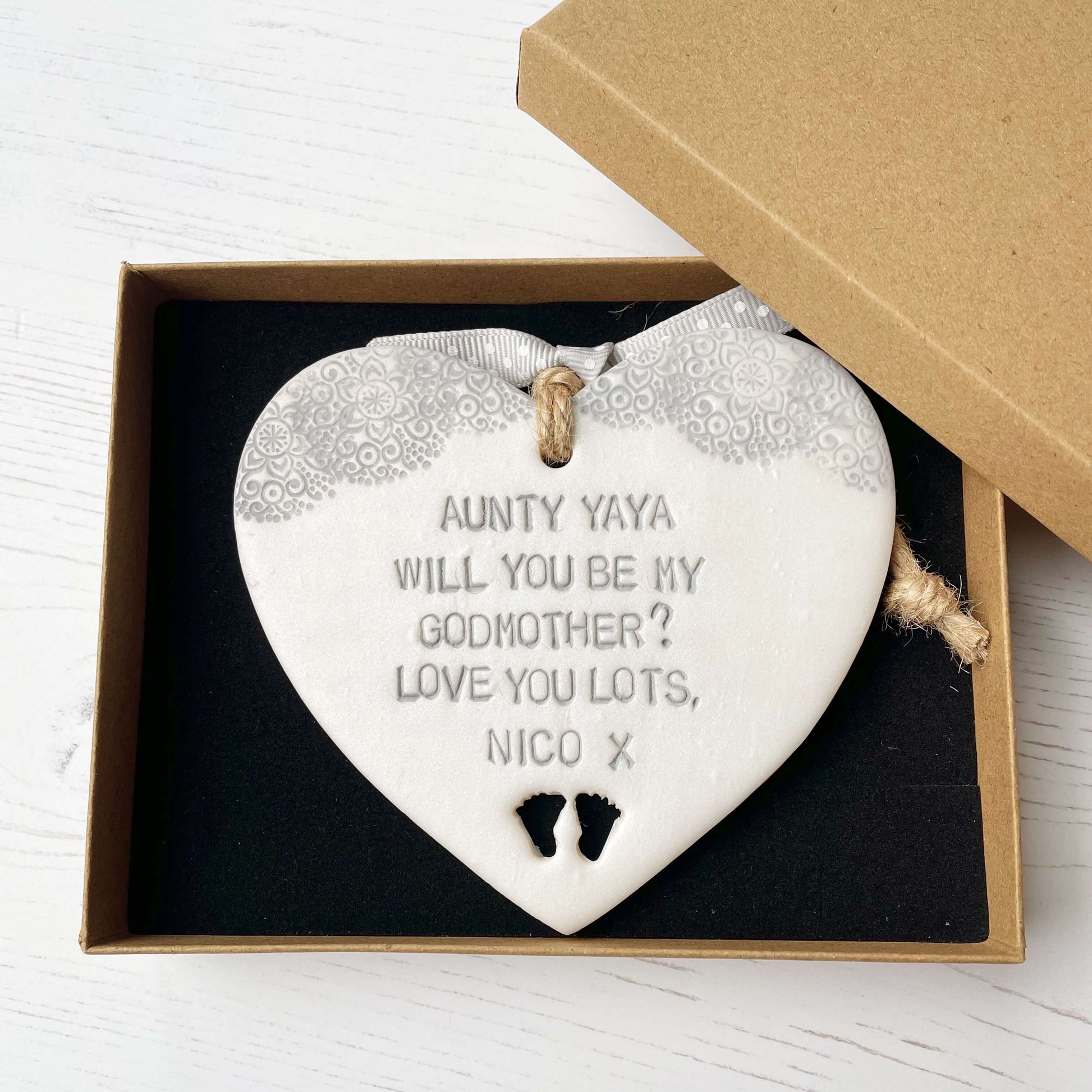 Personalised Godmother gift, pearlised white clay hanging heart with baby feet cut out of the bottom and grey lace edge at the top of the heart, the heart is personalised with AUNTY YAYA, WILL YOU BE MY GODMOTHER? LOVE YOU LOTS NICO X