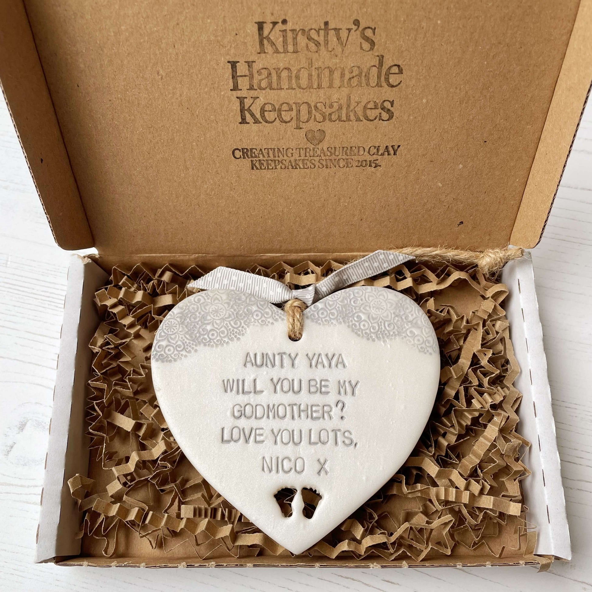 Personalised Godmother gift, pearlised white clay hanging heart with baby feet cut out of the bottom and grey lace edge at the top of the heart, the heart is personalised with AUNTY YAYA, WILL YOU BE MY GODMOTHER? LOVE YOU LOTS NICO X