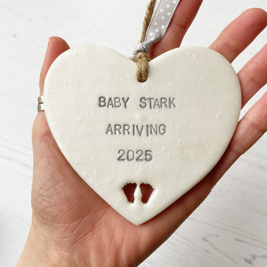 Pearlised white clay hanging heart with baby feet cut out of the bottom and grey personalisation, the heart is personalised with BABY STARK ARRIVING 2025