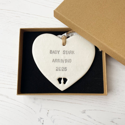 Pearlised white clay hanging heart with baby feet cut out of the bottom and grey personalisation, the heart is personalised with BABY STARK ARRIVING 2025