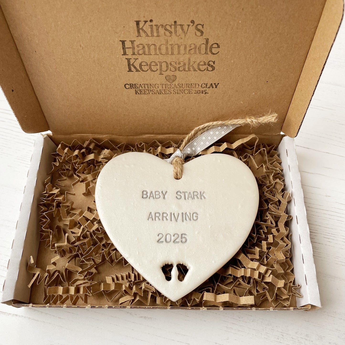 Pearlised white clay hanging heart with baby feet cut out of the bottom and grey personalisation, the heart is personalised with BABY STARK ARRIVING 2025