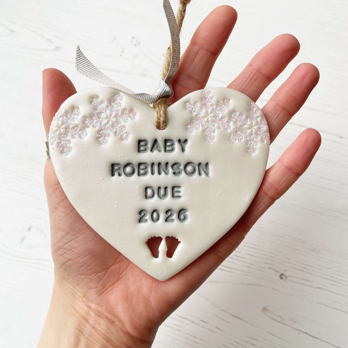 Personalised bumps first Christmas heart ornament, pearlised white clay with BABY ROBINSON DUE 2026 painted grey, decorated with 2 iridescent glitter snowflakes on either side of the top of the heart and baby feet cut out at the bottom