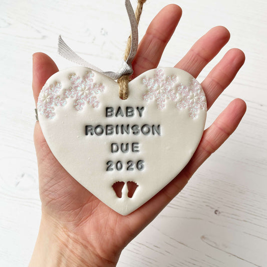 Personalised bumps first Christmas heart ornament, pearlised white clay with BABY ROBINSON DUE 2026 painted grey, decorated with 2 iridescent glitter snowflakes on either side of the top of the heart and baby feet cut out at the bottom