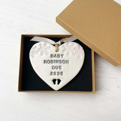 Personalised bumps first Christmas heart ornament, pearlised white clay with BABY ROBINSON DUE 2026 painted grey, decorated with 2 iridescent glitter snowflakes on either side of the top of the heart and baby feet cut out at the bottom