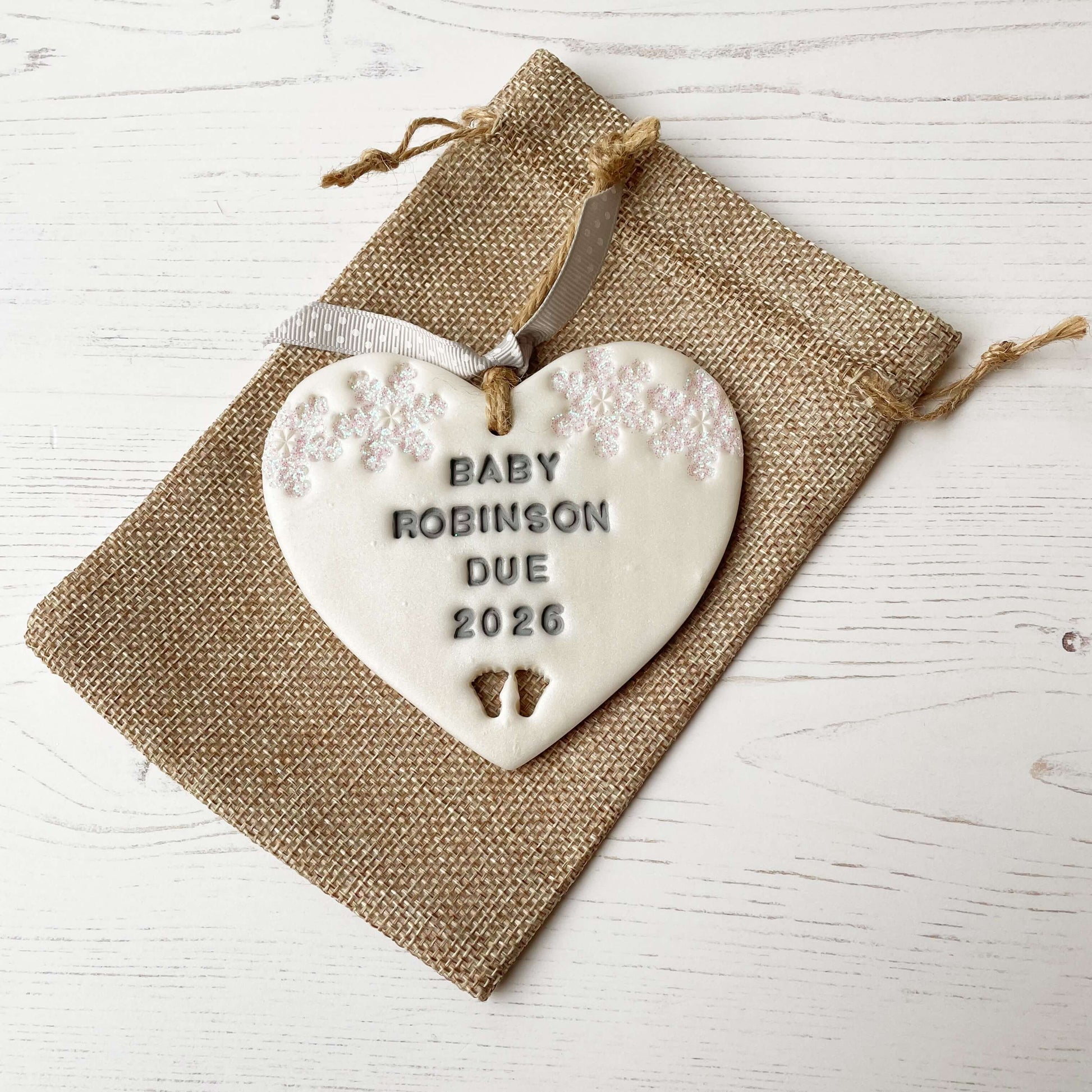 Personalised bumps first Christmas heart ornament, pearlised white clay with BABY ROBINSON DUE 2026 painted grey, decorated with 2 iridescent glitter snowflakes on either side of the top of the heart and baby feet cut out at the bottom